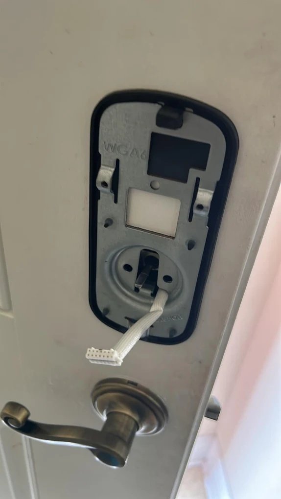 Our Residential Locksmith Merced team working on Close-up of a disassembled smart lock on a white door showing the metal mounting plate and a white ribbon cable with a multi-pin connector. A bronze lever handle is positioned underneath.